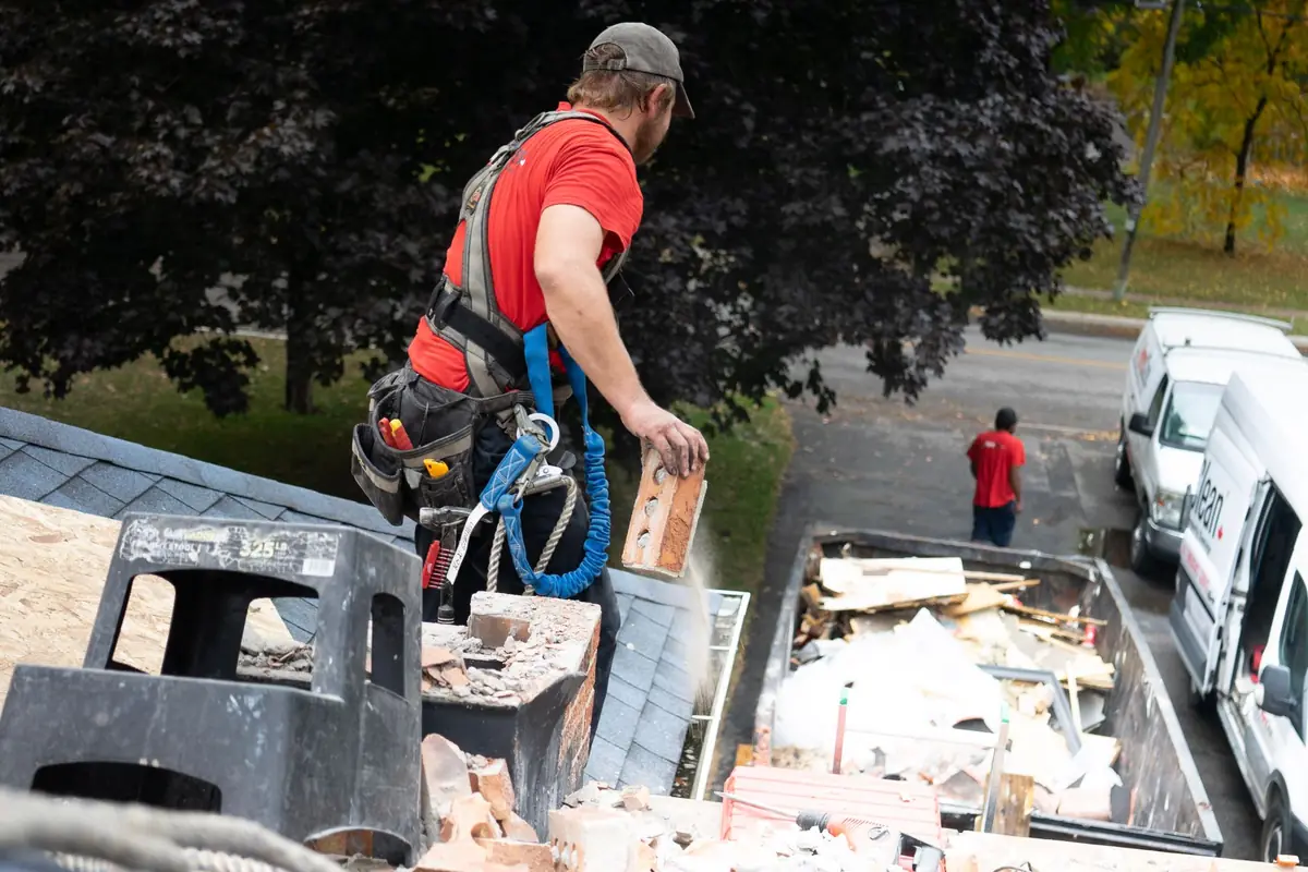 Roofer in a safety harness dropping torn-out chimney brick into a dumpster from a shingle rooftop.