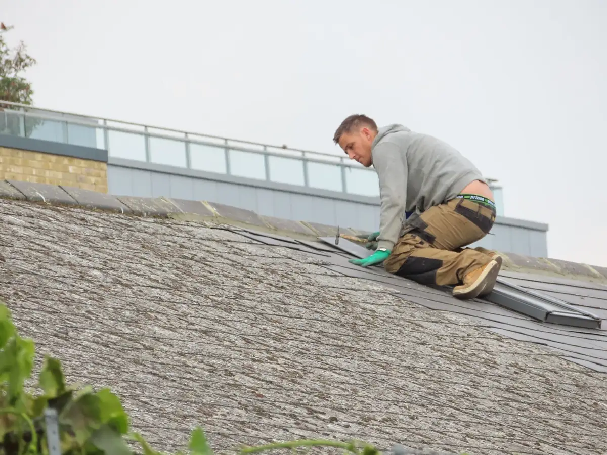 Roofer kneeling near the ridge of a residential roof, fitting a row of dark roof tiles by hand