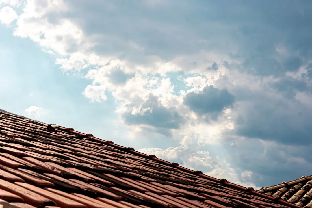 Close-up of a terracotta tile roof against a bright blue sky with scattered clouds.