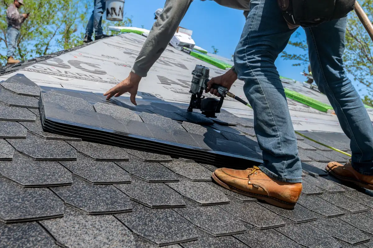 A roofer uses a nail gun to install shingles on a residential roof, the kind of job a yard sign advertises.